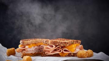 En grillet sandwich med flere lag af kød og ost, serveret med små kartoffeltotter mod en mørk baggrund.