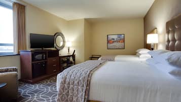 Quarto de hotel com duas camas, uma mesa com televisão, uma cadeira e uma janela grande com cortinas.