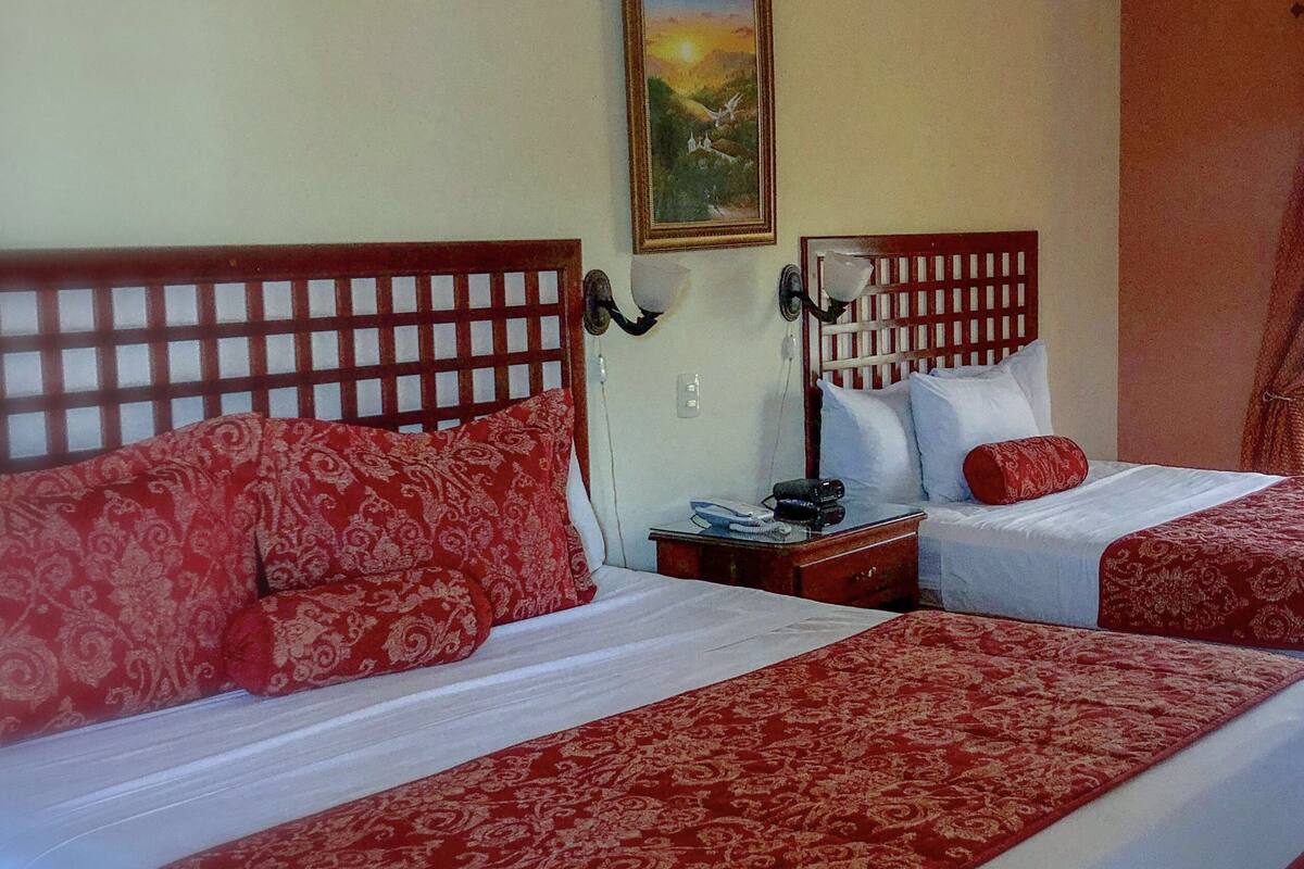 A hotel room with two beds, each with a wooden headboard and red patterned bedspreads. There is a nightstand with a lamp and a telephone between the beds. A painting hangs on the wall above the beds.