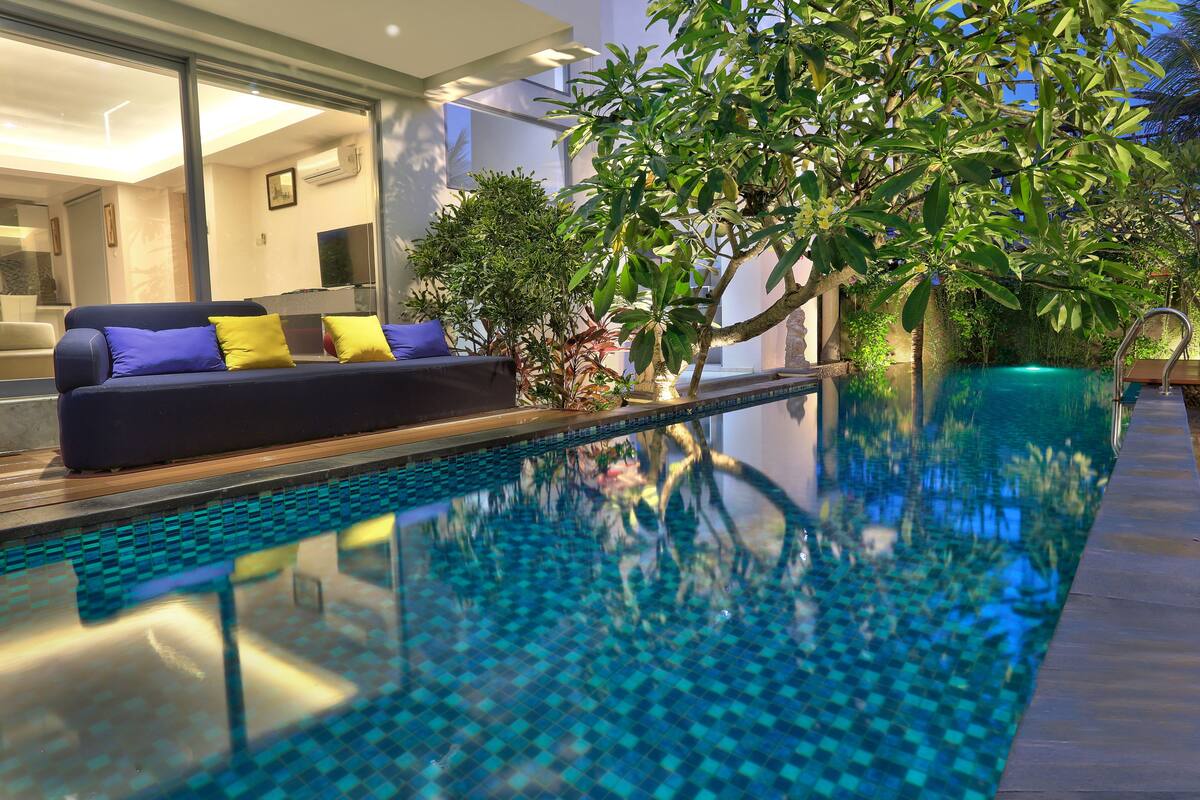 An indoor swimming pool with a tiled floor, surrounded by a wooden deck and a sofa with cushions.