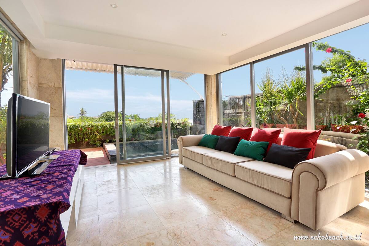 A spacious living room with a beige sofa, a flat-screen TV, and a glass door leading to a balcony with a view of greenery and a clear sky.
