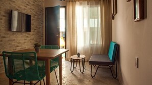 Una habitación con pared de ladrillo, un televisor de pantalla plana, una mesa de comedor con sillas, una pequeña mesa redonda con una planta, un banco y una ventana con cortinas traslúcidas.
