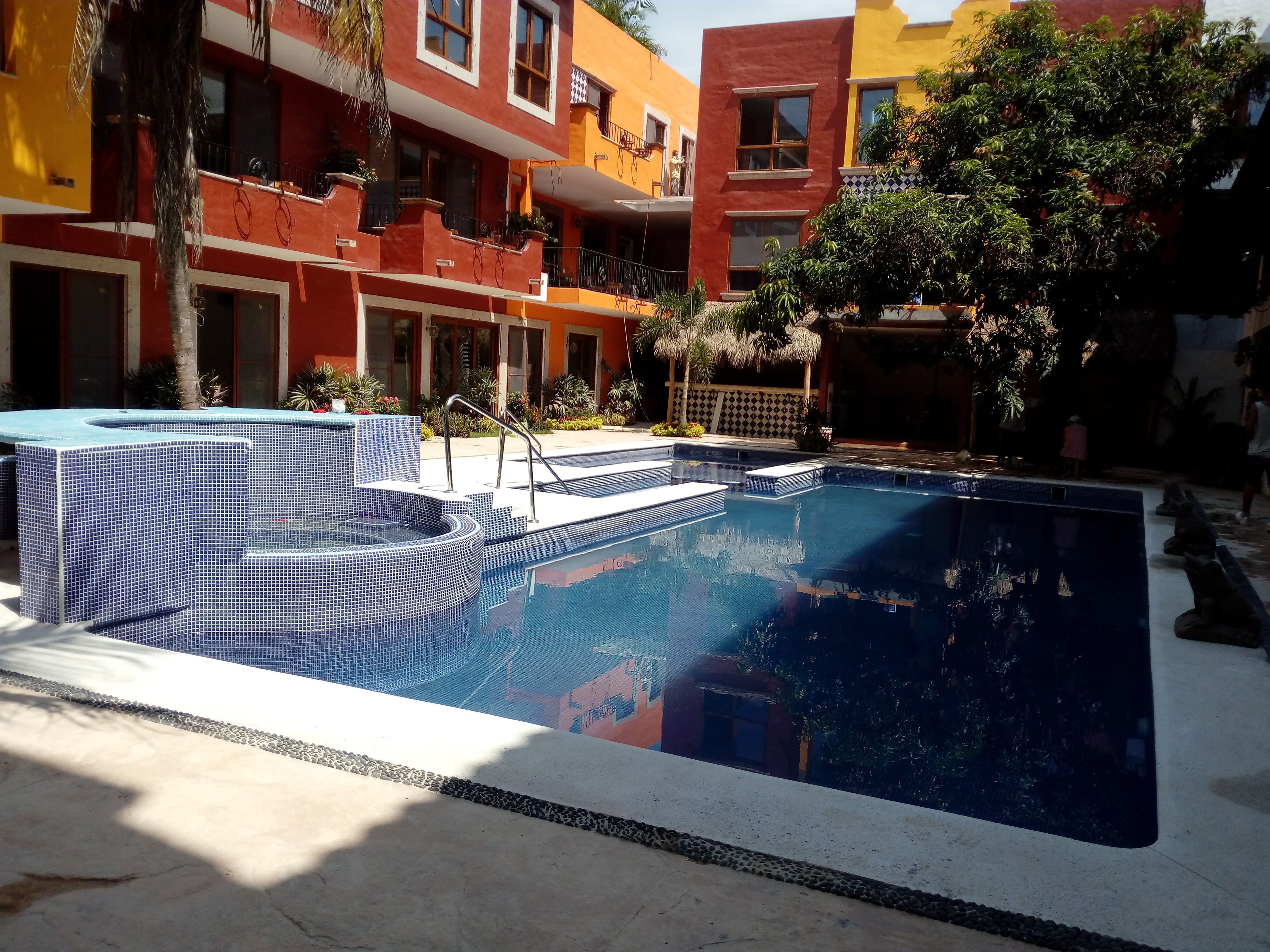 A swimming pool with tiled steps and a hot tub, surrounded by colorful buildings and greenery.