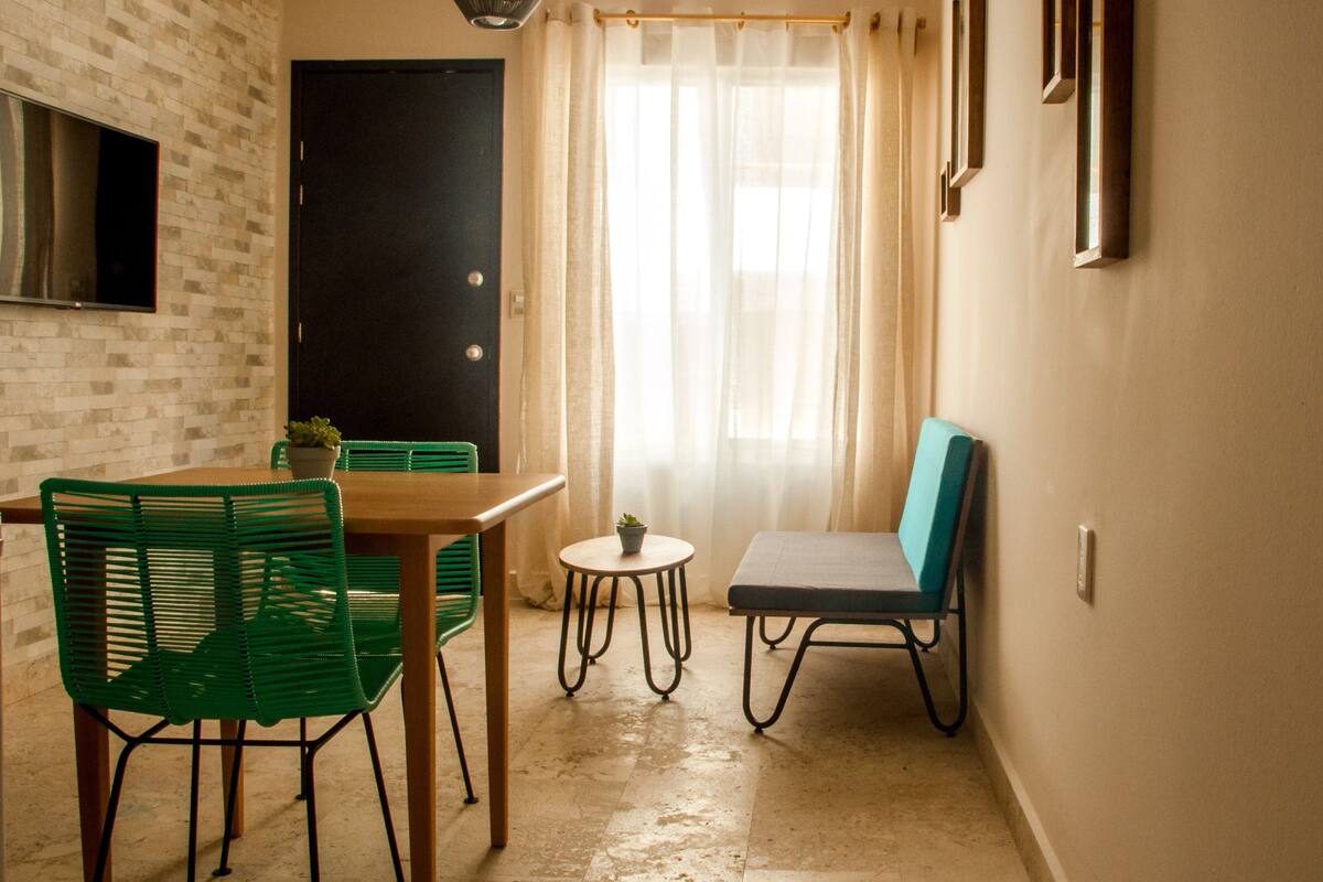 Un petit coin repas avec une table et des chaises, une plante en pot et une vue sur une porte et une fenêtre avec des rideaux.