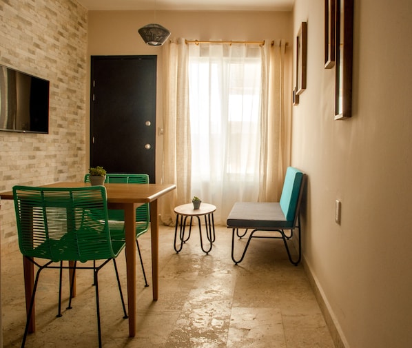 Un pequeño comedor con una mesa y sillas, una planta en maceta y vistas de una puerta y una ventana con cortinas.