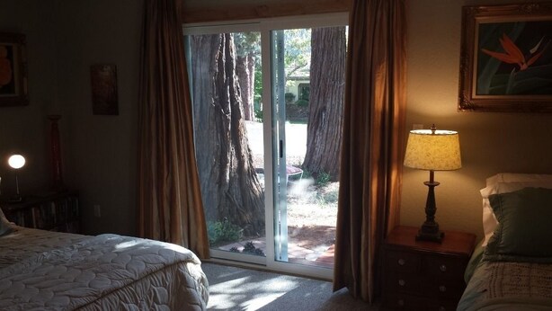 Un dormitorio con una cama, una mesita de noche con lámpara y un ventanal corredero que da a un espacio exterior con árboles.