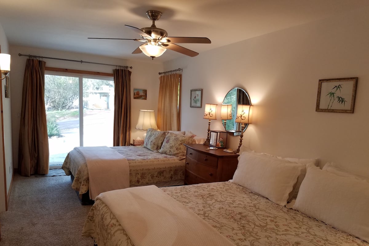 A bedroom with two beds, a ceiling fan, a dresser with lamps, and a window with curtains.