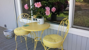 Un conjunto de mesa y sillas de metal amarillo sobre una terraza de madera, con una maceta de rosas rosadas en el alféizar.
