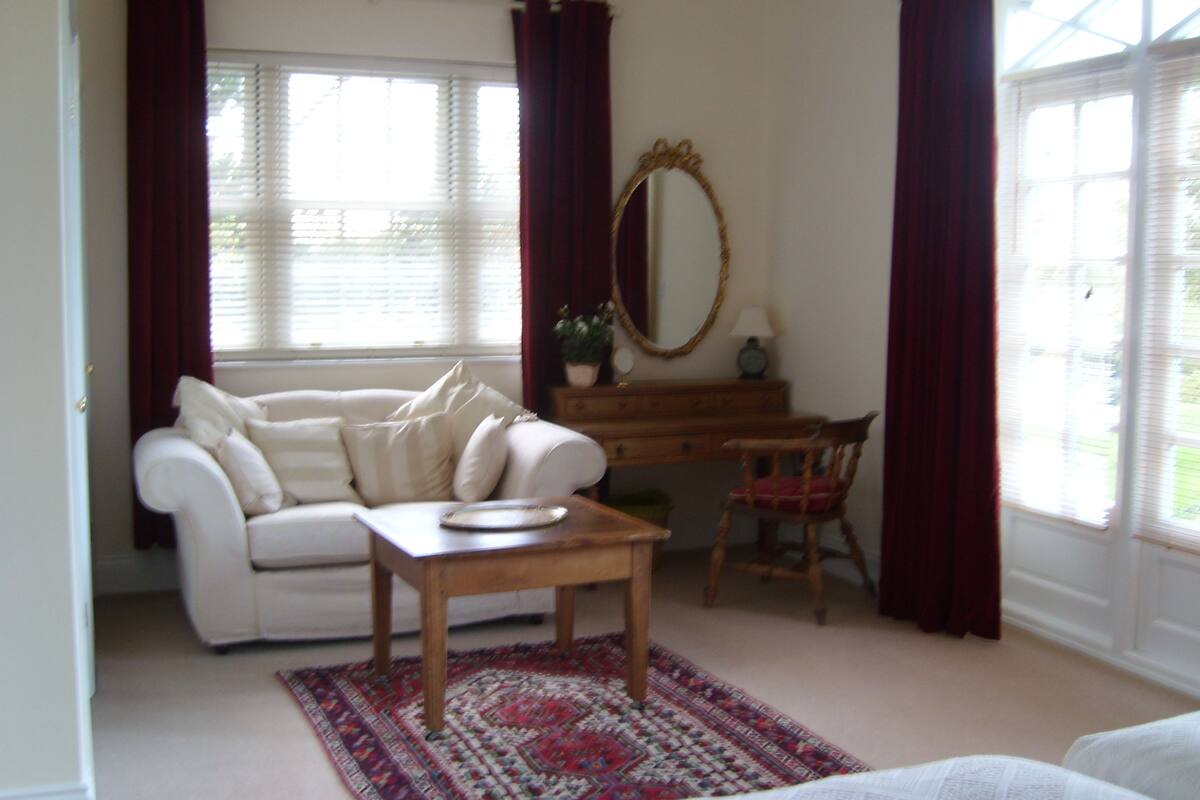 Une pièce avec un canapé blanc, une table en bois, un bureau et une chaise. Il y a un miroir au mur et une fenêtre avec des rideaux.