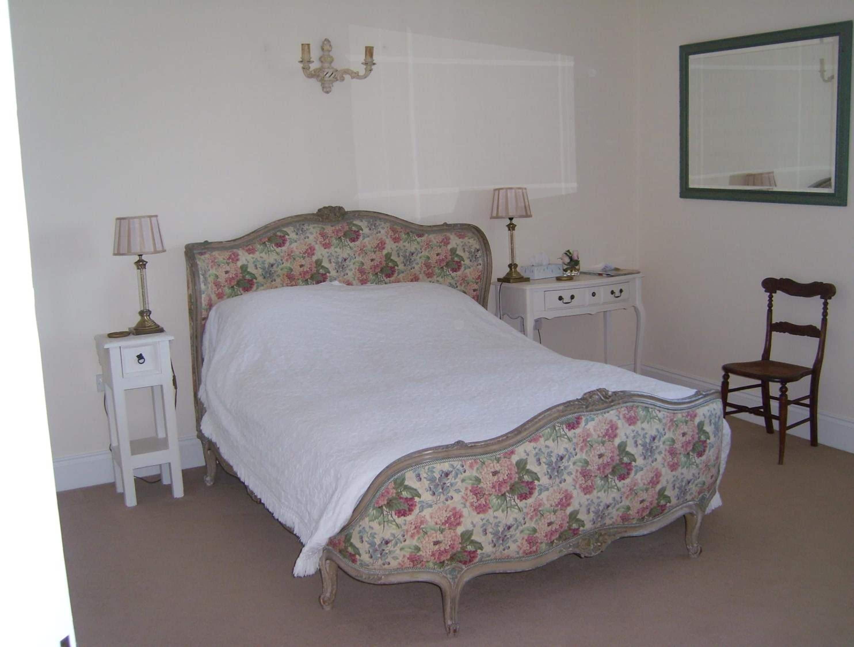A bedroom with a floral-patterned bed, two bedside tables with lamps, a chair, and a mirror on the wall.