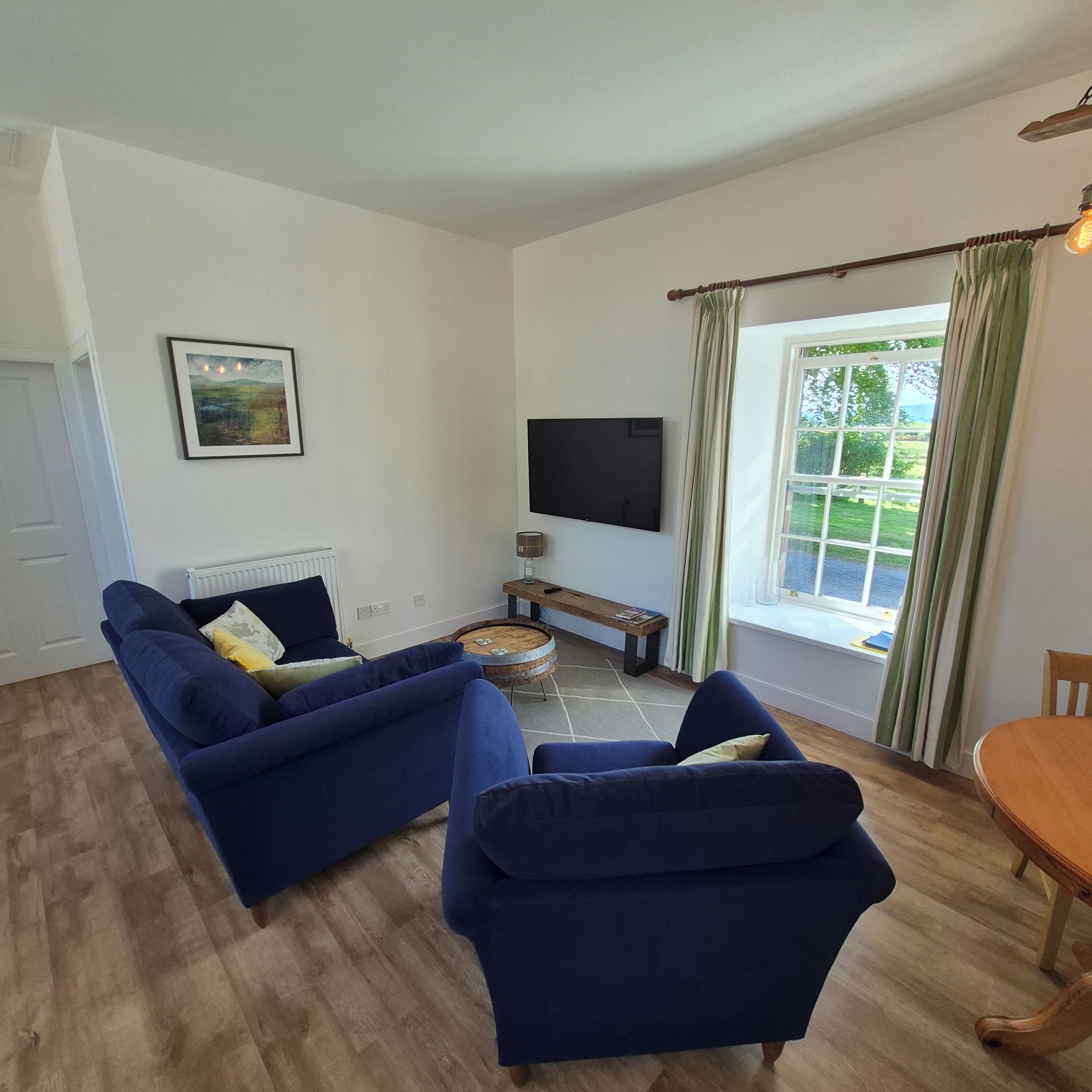 A living room with a blue sofa set, a flat-screen TV, a round wooden coffee table, and a window with curtains.