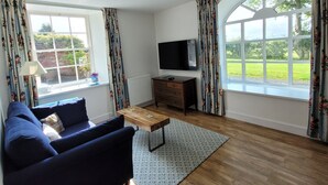 Sala de estar com sofá, mesa de centro, televisão e vista para um jardim por grandes janelas.