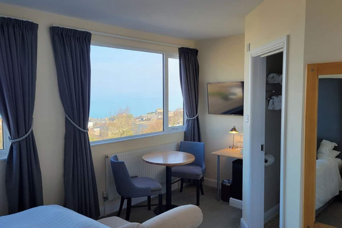 A hotel room with a bed, a small table with two chairs, a view of the sea, and a closet with clothes.