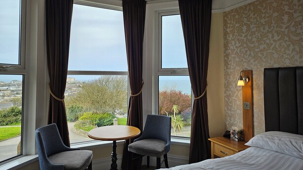 A hotel room with a bed, a small table, two chairs, a nightstand, and a lamp. There are large windows with curtains and a view of the outside. - Geckos Rest (Newquay)