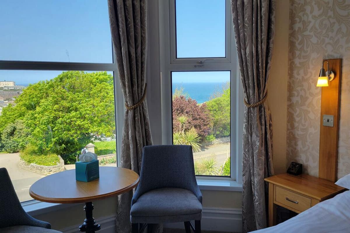 A hotel room with a bed, a table, a chair, a nightstand, and a view of the sea and greenery.