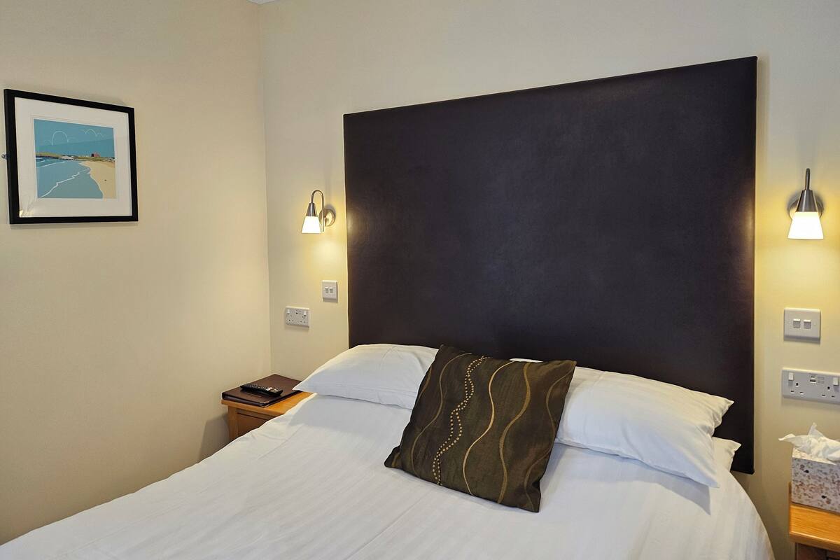 A hotel room with a large bed, two bedside tables, a nightstand, a framed picture on the wall, and two wall-mounted lights.
