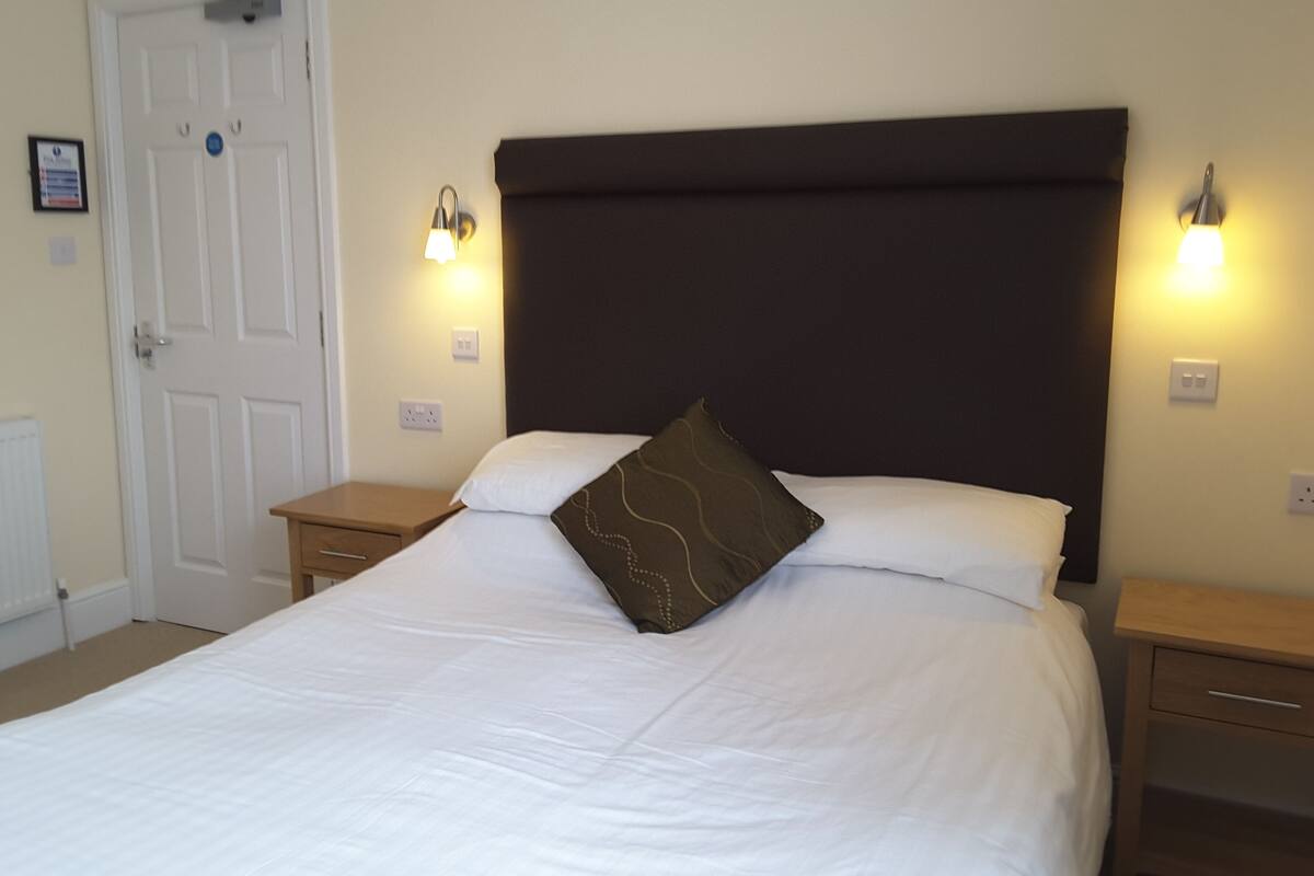 A neatly made bed with white linens and a brown decorative pillow, flanked by two bedside tables with lamps, in a room with a white door and a light switch on the wall.