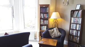 A living room with a blue armchair, a small wooden table, and a bookshelf filled with books and DVDs. - Geckos Rest (Newquay)
