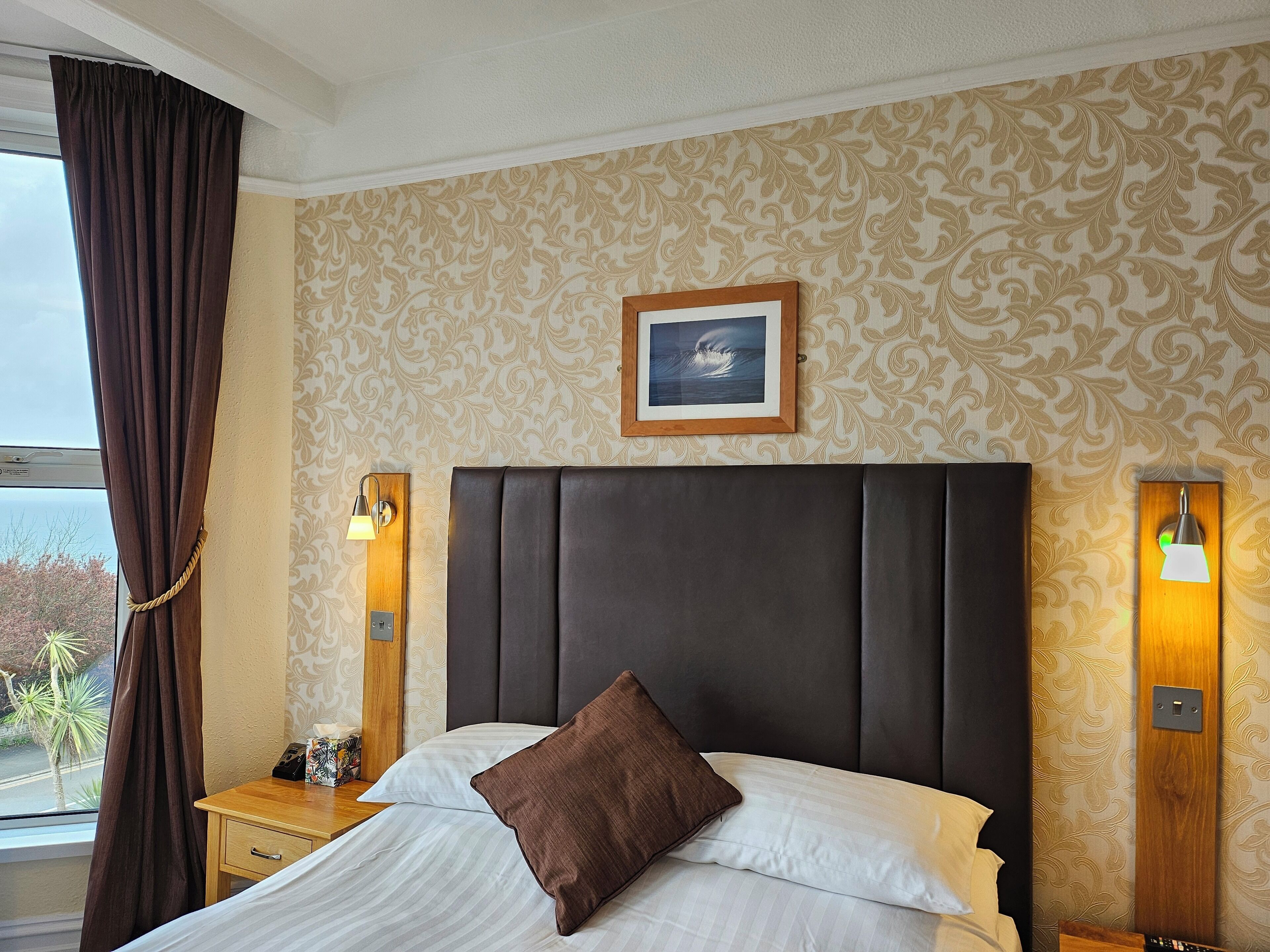 A hotel room with a large bed, a headboard, two bedside lamps, a nightstand, a framed picture on the wall, and a window with curtains.