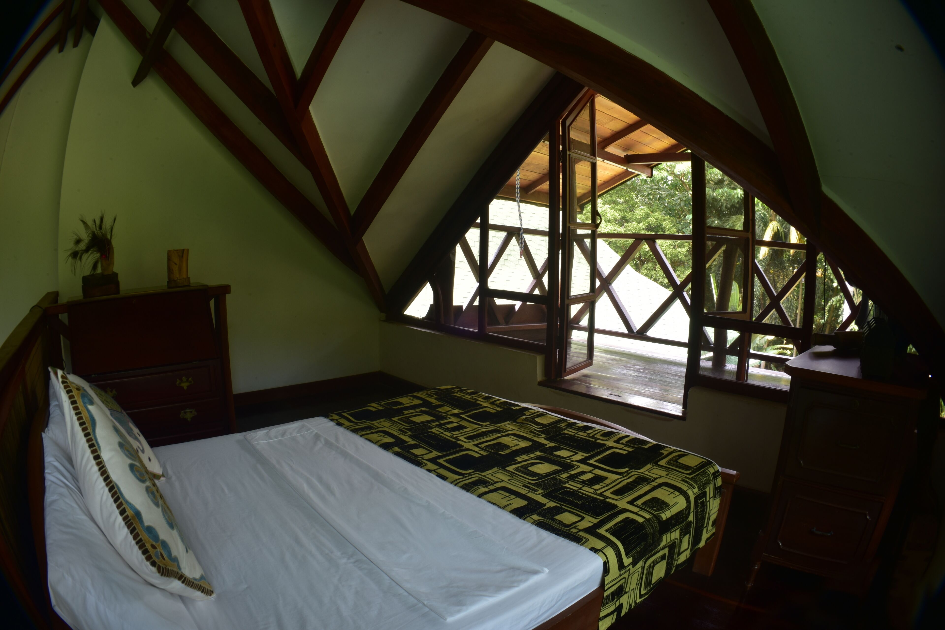 Ein Schlafzimmer mit schrägem Dach, einem Bett mit gemustertem Überwurf, einem Nachttisch, einer Kommode und einem Fenster mit Blick auf Grünflächen.