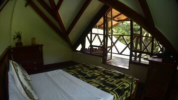Ein Schlafzimmer mit schrägem Dach, einem Bett mit gemustertem Überwurf, einem Nachttisch, einer Kommode und einem Fenster mit Blick auf Grünflächen.