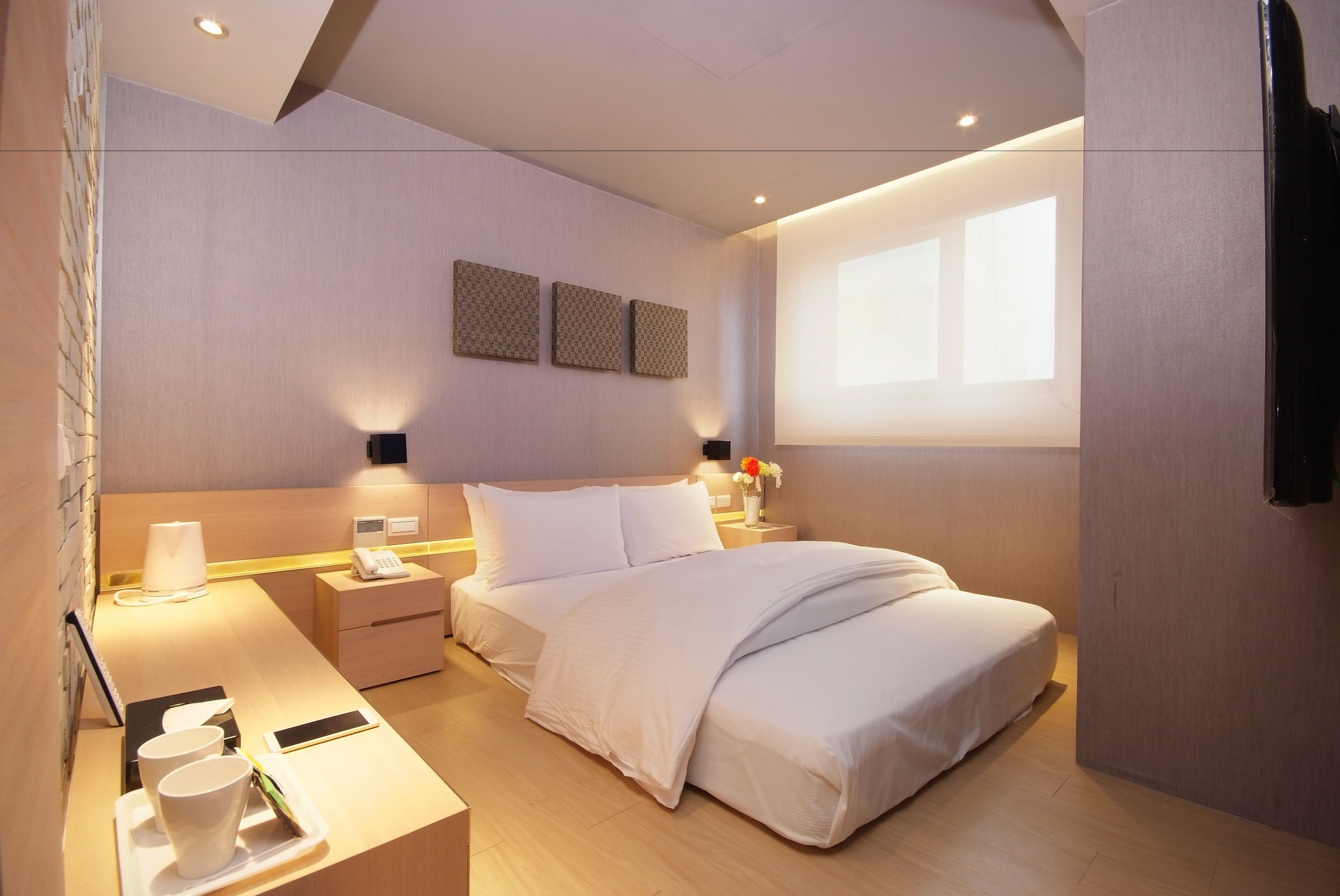 A modern hotel room with a neatly made bed, bedside tables with lamps, a small desk with a cup and a phone, and a large window providing natural light.