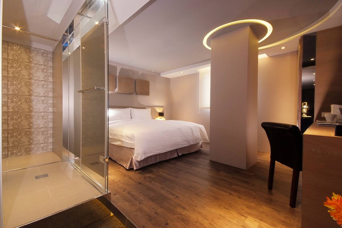 A modern hotel room with a glass-enclosed shower, a neatly made bed, a wooden floor, and a small desk area with a chair.