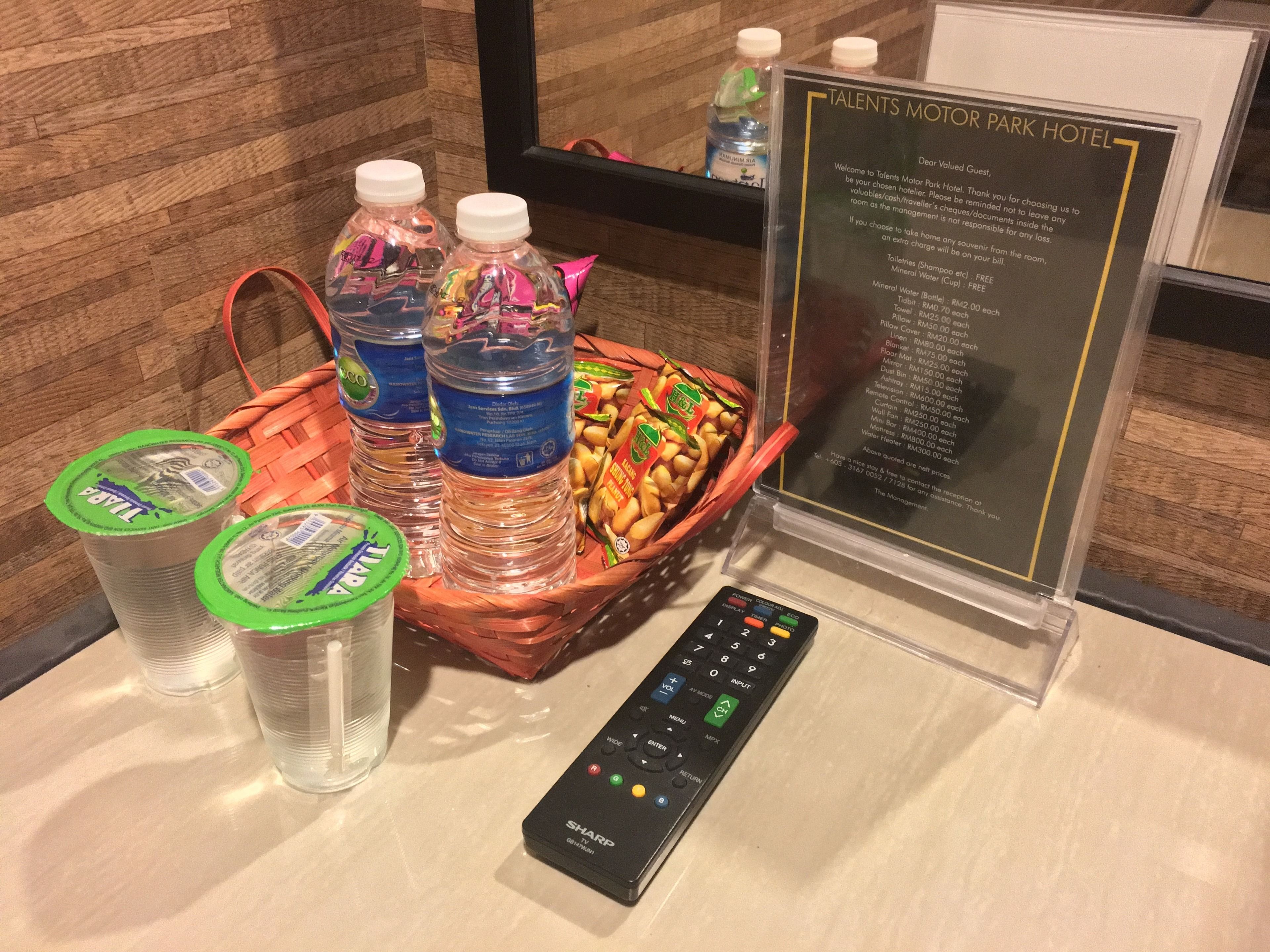 Un espace d’accueil d’hôtel avec un panneau indiquant les services proposés et les tarifs, un panier contenant des bouteilles d’eau et des en-cas, et une télécommande.
