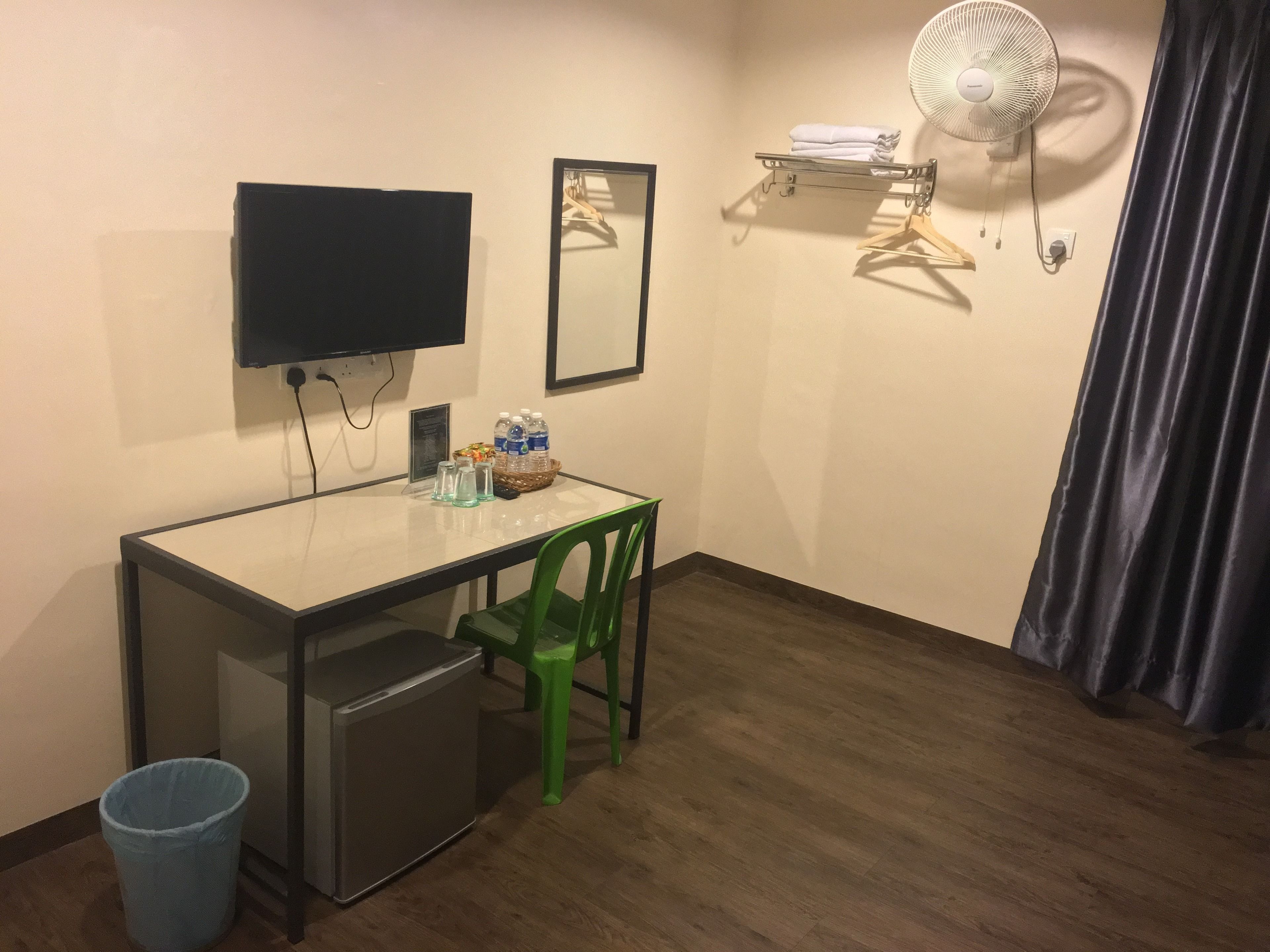 A small room with a desk, a green chair, a small fridge, a trash bin, a television, a mirror, a fan, and a shelf with folded towels.