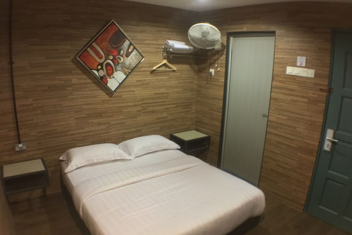 Un dormitorio con una cama, una mesita de noche, un ventilador de pared, una puerta y un cuadro enmarcado en la pared.