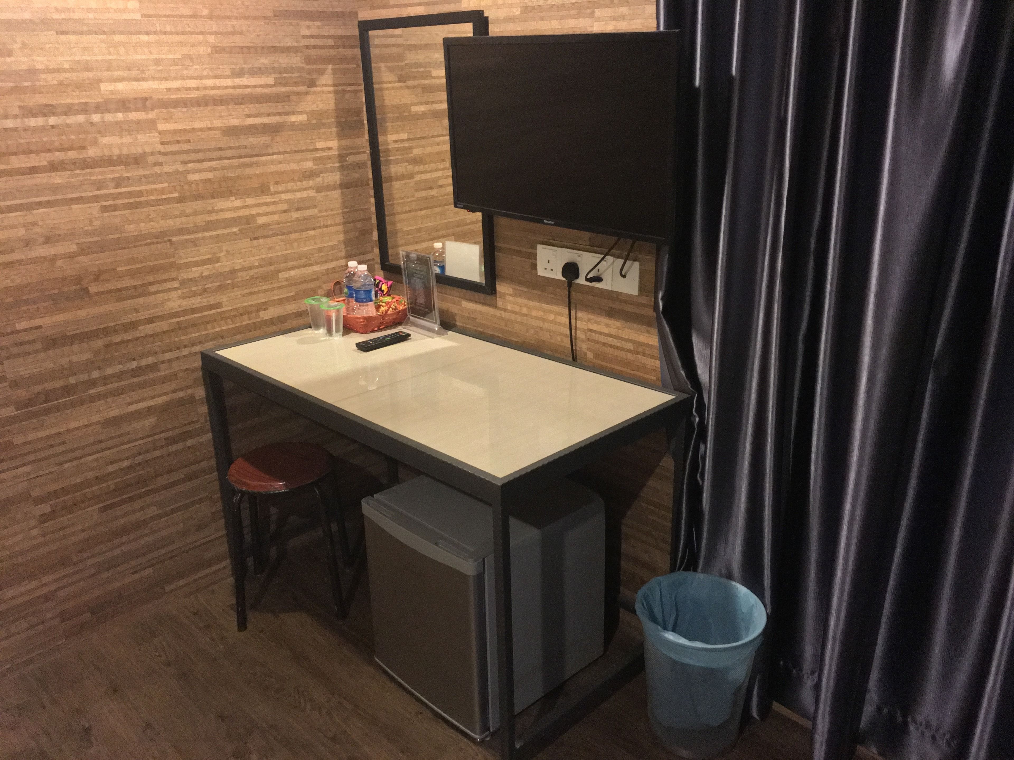 A compact room with a small desk, a television mounted on the wall, a trash bin, and a compact refrigerator.