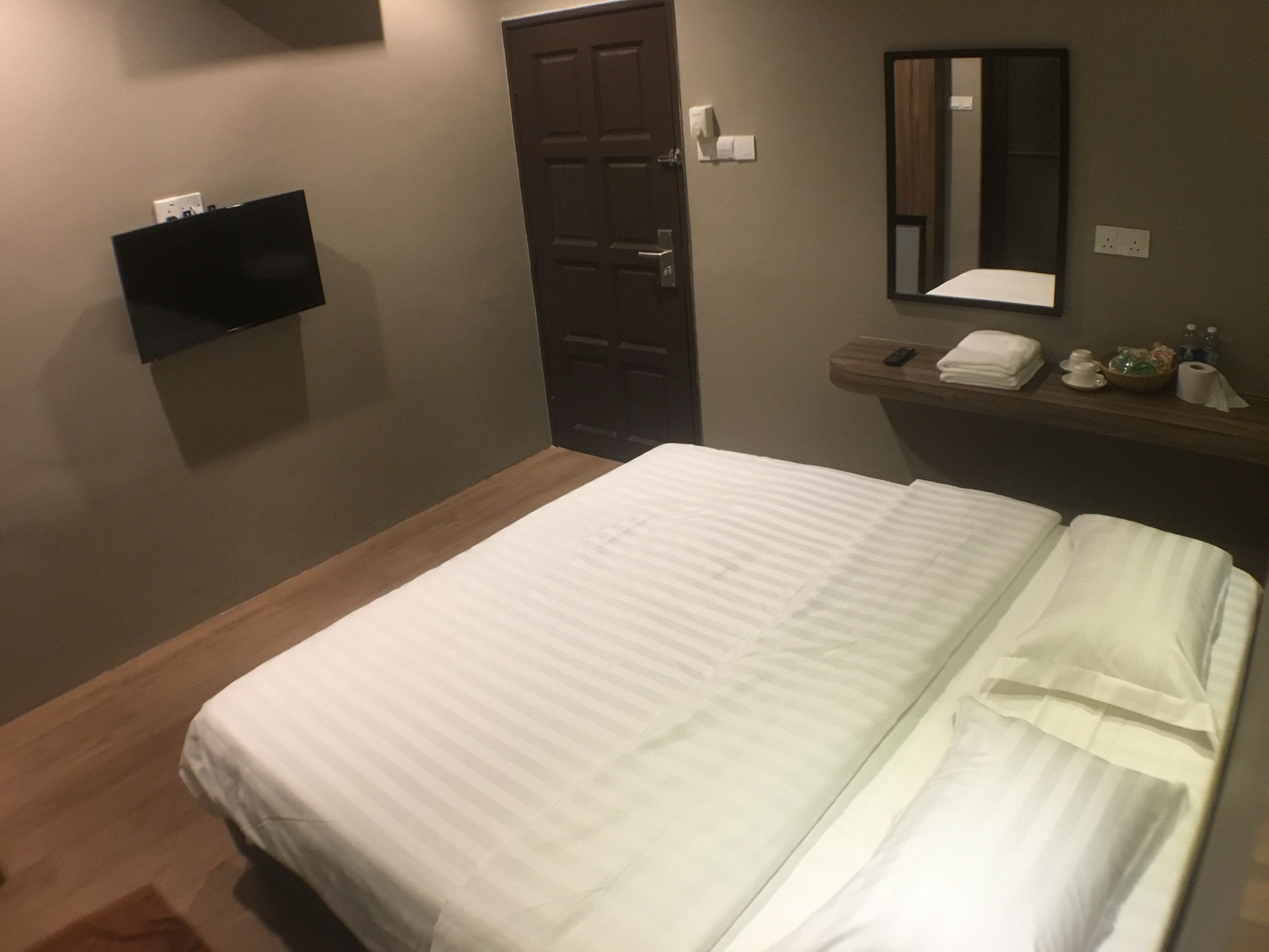 A hotel room with a bed, a wooden floor, a door, a mirror, and a shelf with toiletries.