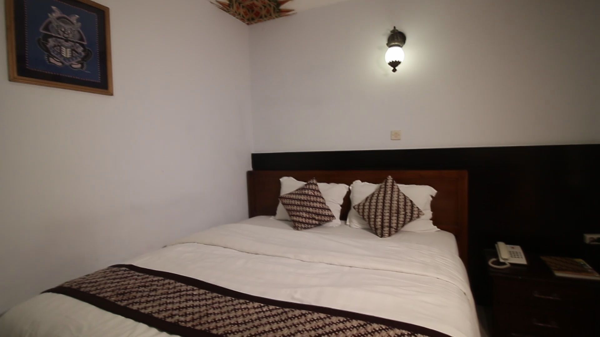 A neatly made bed with a patterned bedspread and pillows, a wooden headboard, a wall-mounted light fixture, and a framed picture on the wall.
