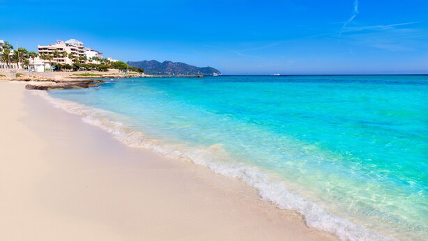 A beach with clear turquoise water, a sandy shore, and a building with multiple floors and balconies in the background. - Hotel Ilusion Vista Blava (Son Servera)