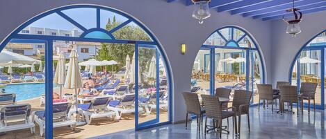 A sunlit indoor area with a pool view, featuring lounge chairs, tables, and arched windows.