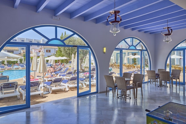 A sunlit indoor area with a pool view, featuring lounge chairs, tables, and arched windows.