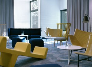 A modern lounge area with a sofa, chairs, and tables, featuring a geometric patterned wall and large windows. - The Jangle Hotel Paris Charles de Gaulle Airport (Le Mesnil-Amelot)