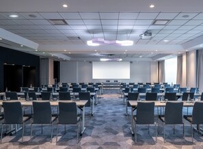 A conference room with rows of tables and chairs, a large screen, and a projector. - The Jangle Hotel Paris Charles de Gaulle Airport (Le Mesnil-Amelot)