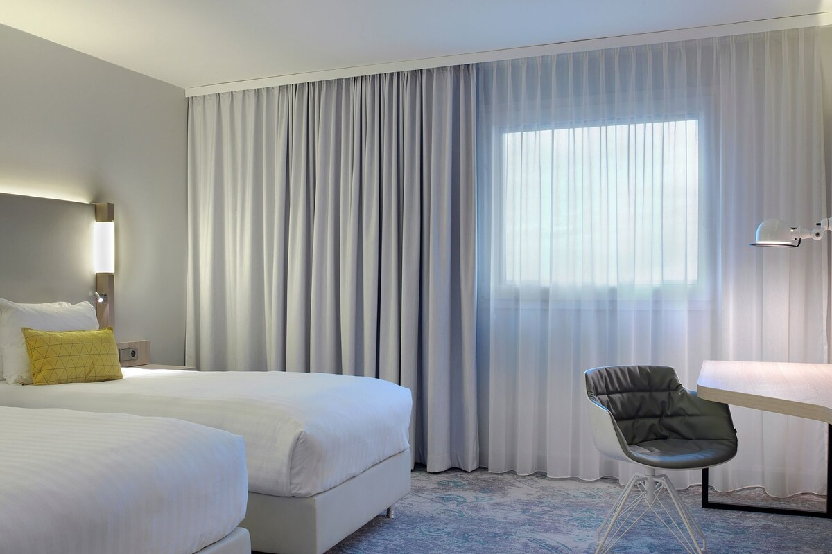 A hotel room with two beds, a desk, a chair, and a large window with sheer curtains.