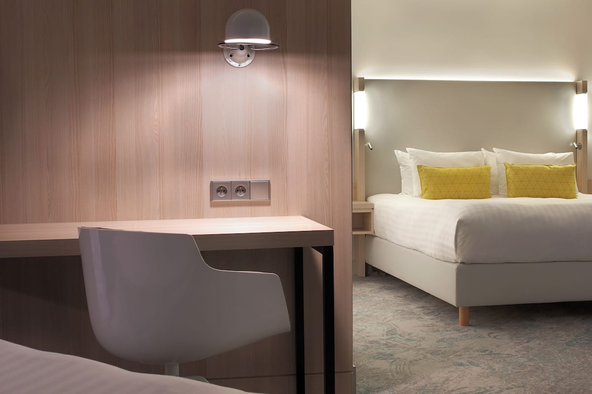 Une chambre d’hôtel avec un mur en bois, un bureau avec une chaise, un lit avec une literie blanche et jaune, et une moquette à motifs.