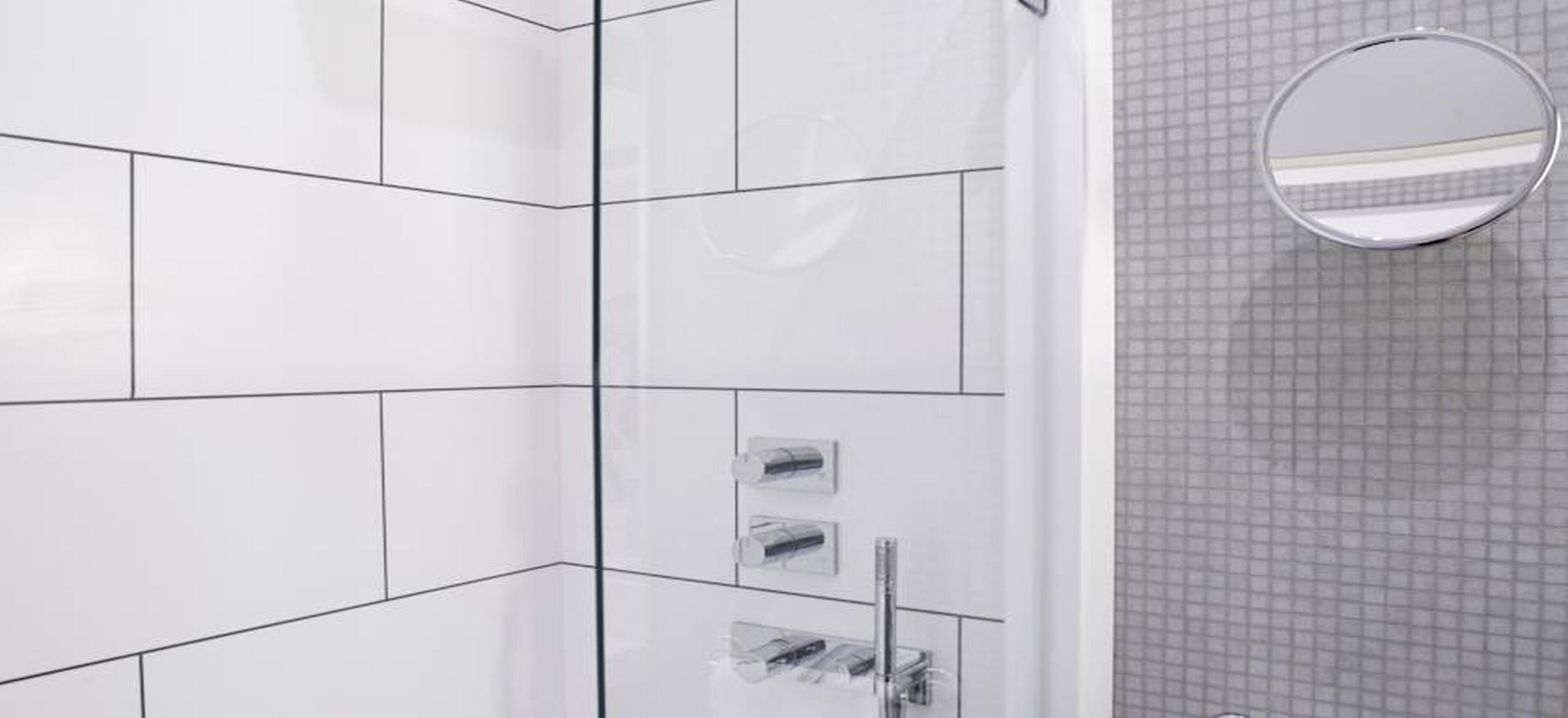 A modern bathroom with white tiles, a glass shower enclosure, and a small round mirror.