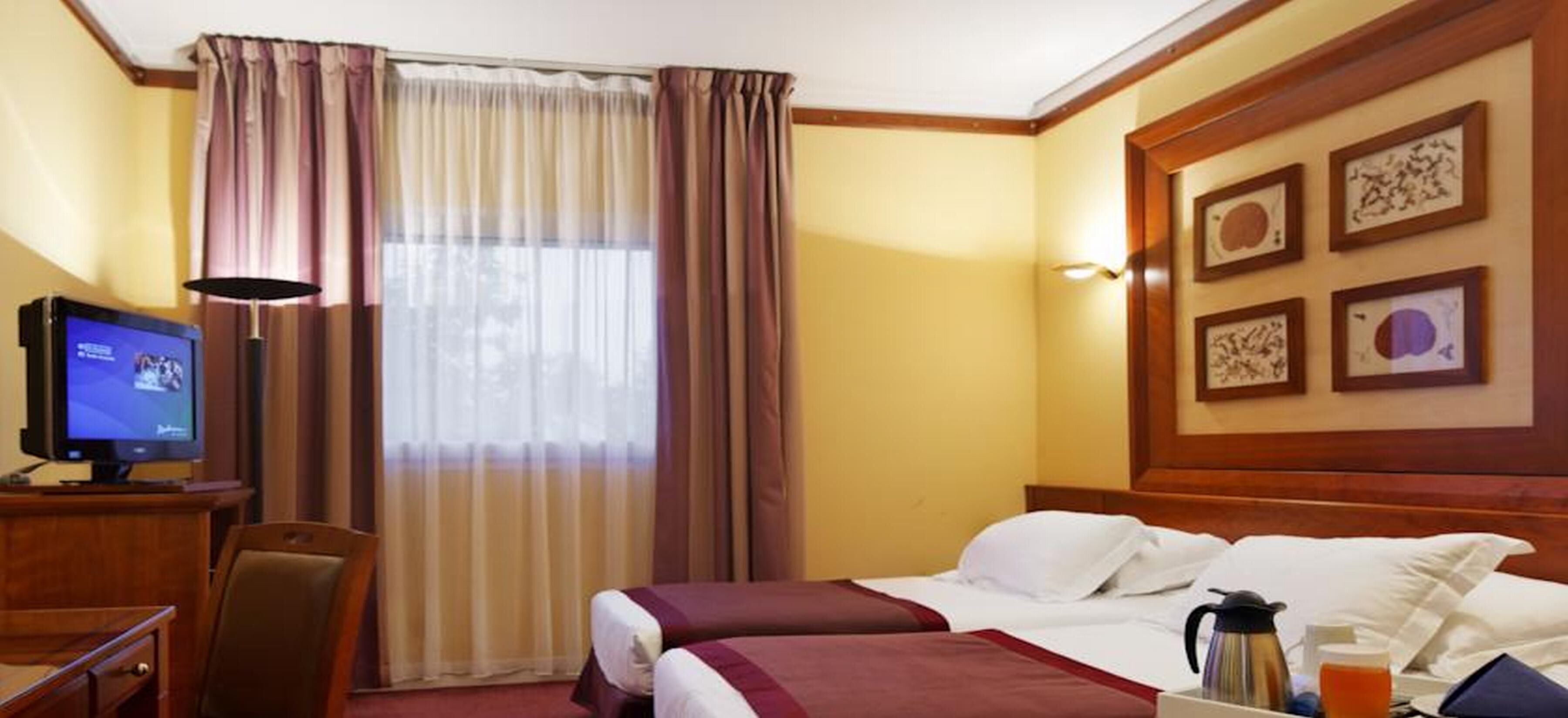 A hotel room with a bed, a desk with a computer, a chair, a window with curtains, and framed artwork on the wall.