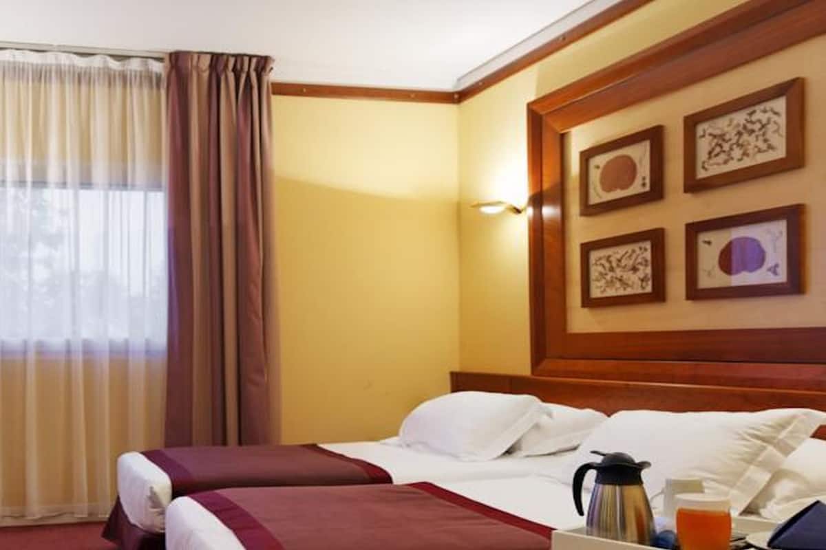 A hotel room with a bed, a desk with a computer, a chair, a window with curtains, and framed artwork on the wall.