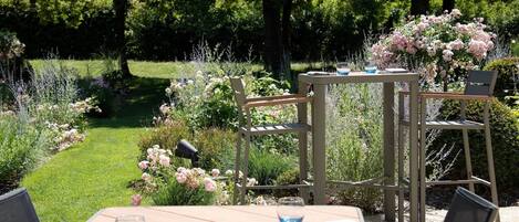 Un área de comedor al aire libre con una mesa y sillas de madera, rodeada de abundante vegetación y plantas con flores.