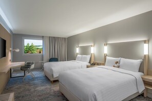 Ein Hotelzimmer mit zwei Betten, einem Schreibtisch, einem Stuhl, einem Fernseher und einem Fenster mit Blick auf Grünflächen.