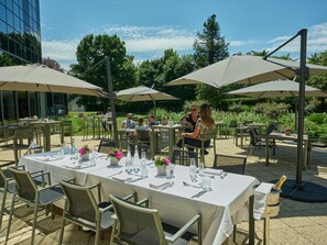 An outdoor dining area with tables and chairs, umbrellas, and a view of greenery and a building in the background. - The Jangle Hotel Paris Charles de Gaulle Airport (Le Mesnil-Amelot)