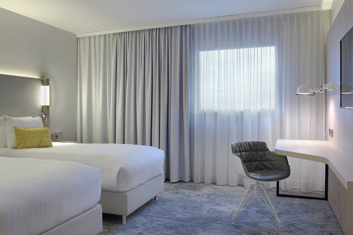 Une chambre d’hôtel avec deux lits, un bureau, une chaise et une grande fenêtre avec des rideaux.