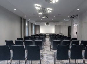 A conference room with rows of chairs, a projector screen, and a ceiling with hexagonal light fixtures. - The Jangle Hotel Paris Charles de Gaulle Airport (Le Mesnil-Amelot)