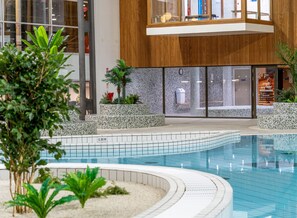 A modern indoor swimming pool with tiled edges, surrounded by planters and a wooden structure with glass windows. - The Jangle Hotel Paris Charles de Gaulle Airport (Le Mesnil-Amelot)