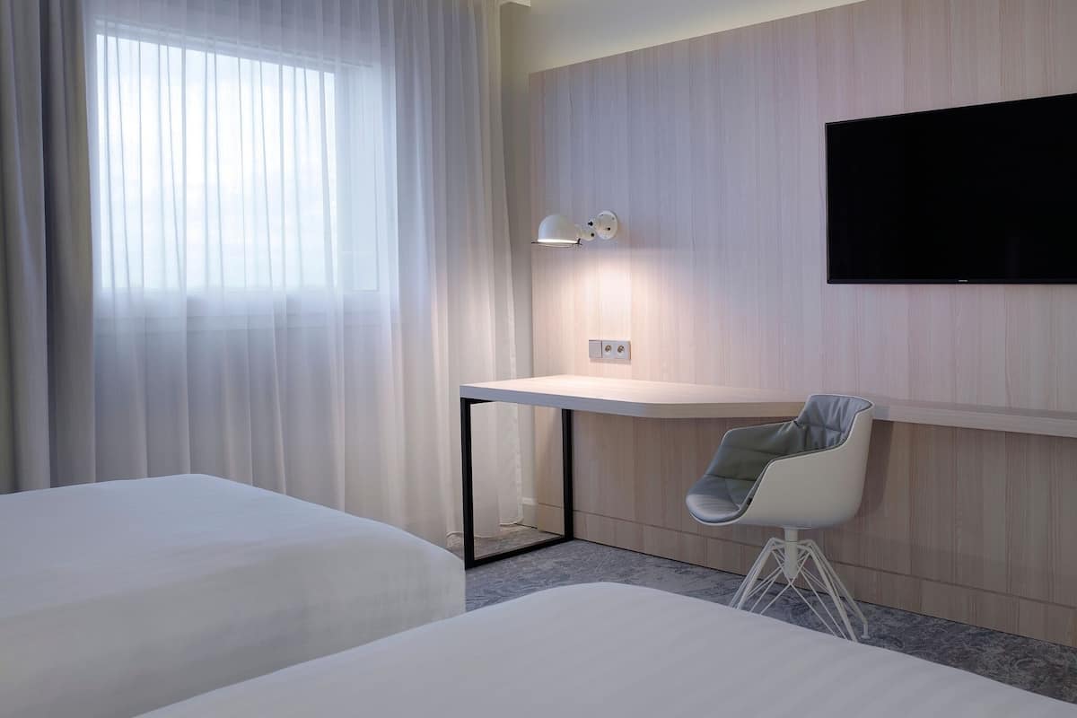 Une chambre d’hôtel avec deux lits, un bureau avec une chaise, un téléviseur fixé au mur et une fenêtre avec des rideaux légers.